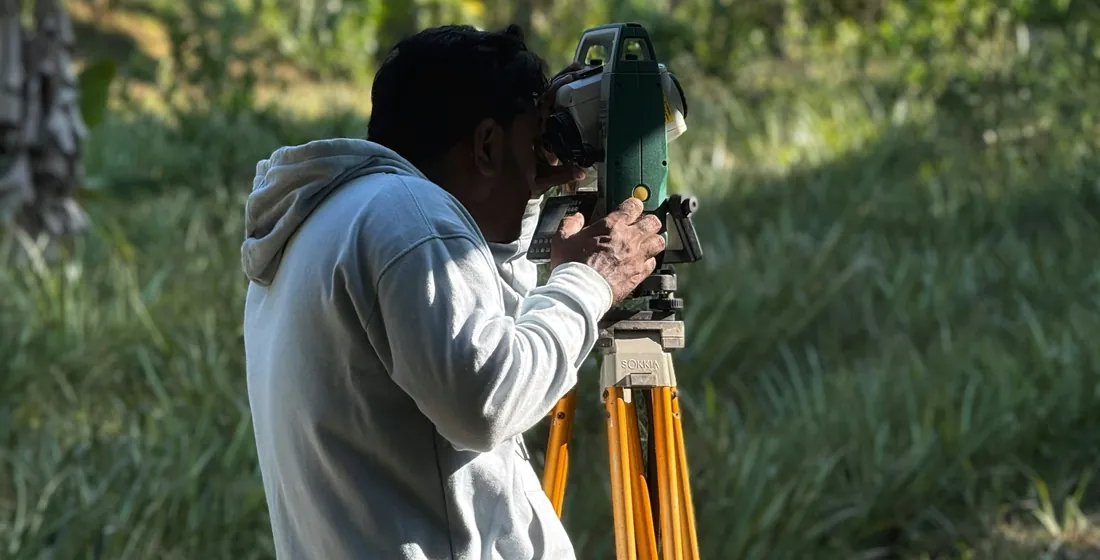 Surveyor working with a total station on a tripod in long grass at the edge of a treeline
