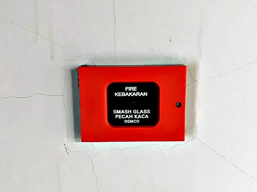Red manual call point marked FIRE, KEBAKARAN, SMASH GLASS, PECAH KACA, mounted on a plain wall