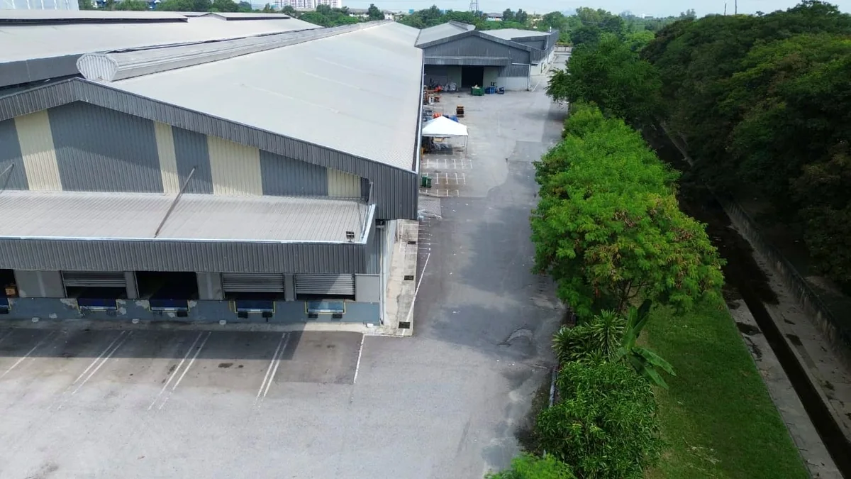 Aerial view of a detached factory with loading docks along one elevation, a wide concrete apron in front of it, and a drainage reserve along the boundary