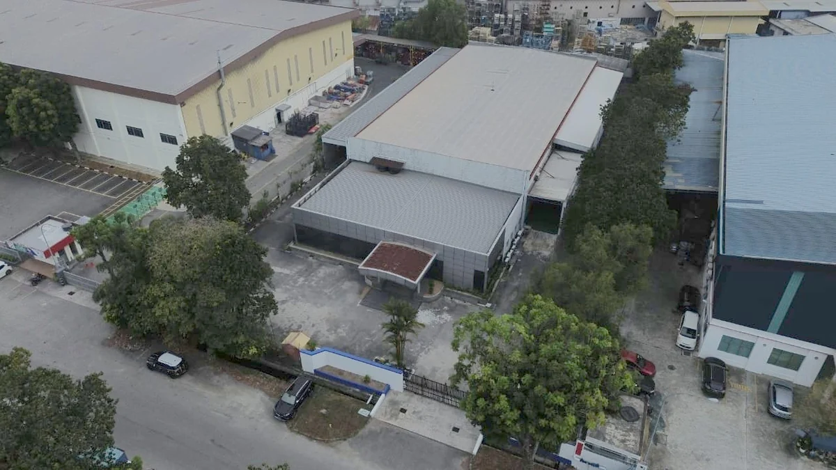 Aerial view of a detached industrial unit seen from the front, with a two storey front block, a canopy over the entrance, an open concrete forecourt with vehicles parked on it, and a boundary wall and gate onto the road