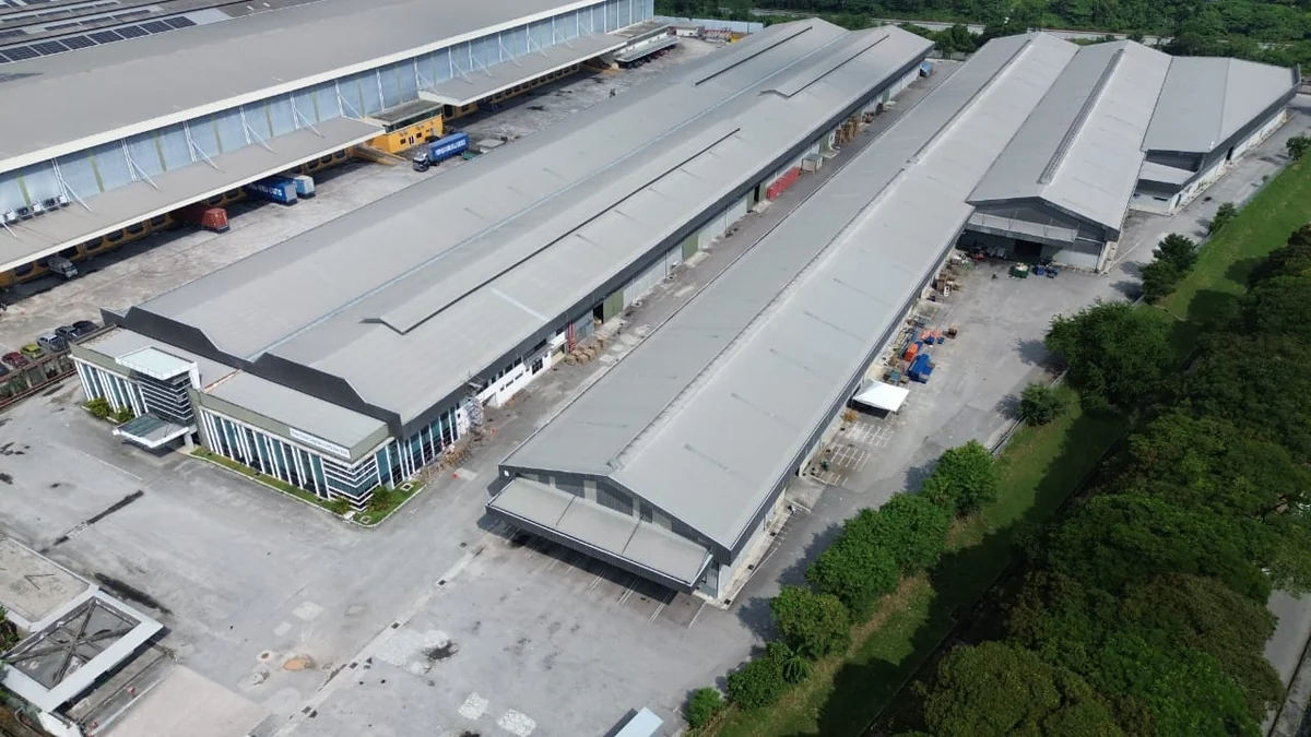 Aerial view of three long parallel industrial spans under one roofline, with a glazed office block at the near corner, a covered canopy over the entrance and a wide concrete apron running the length of the elevation