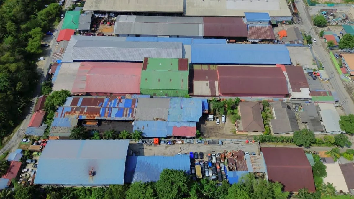 Rooflines of an older industrial estate photographed from above, weathered metal sheeting with patch repairs, ventilators along the ridges and mature trees along the boundary