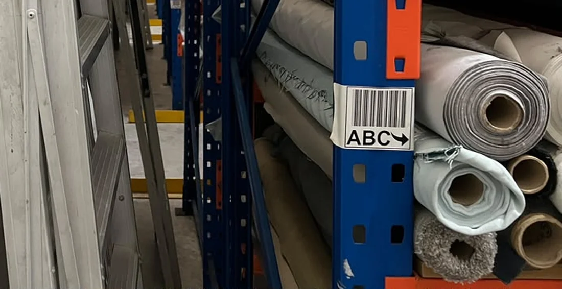 Pallet racking uprights in a warehouse aisle, carrying rolled stock on the beams